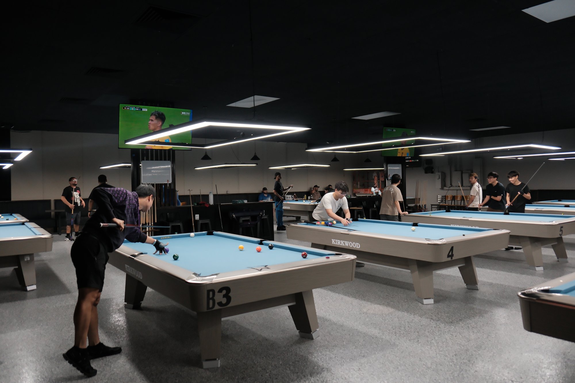 Players in action on Kirkwood Billiards tournament-grade tables with sports on the TVs